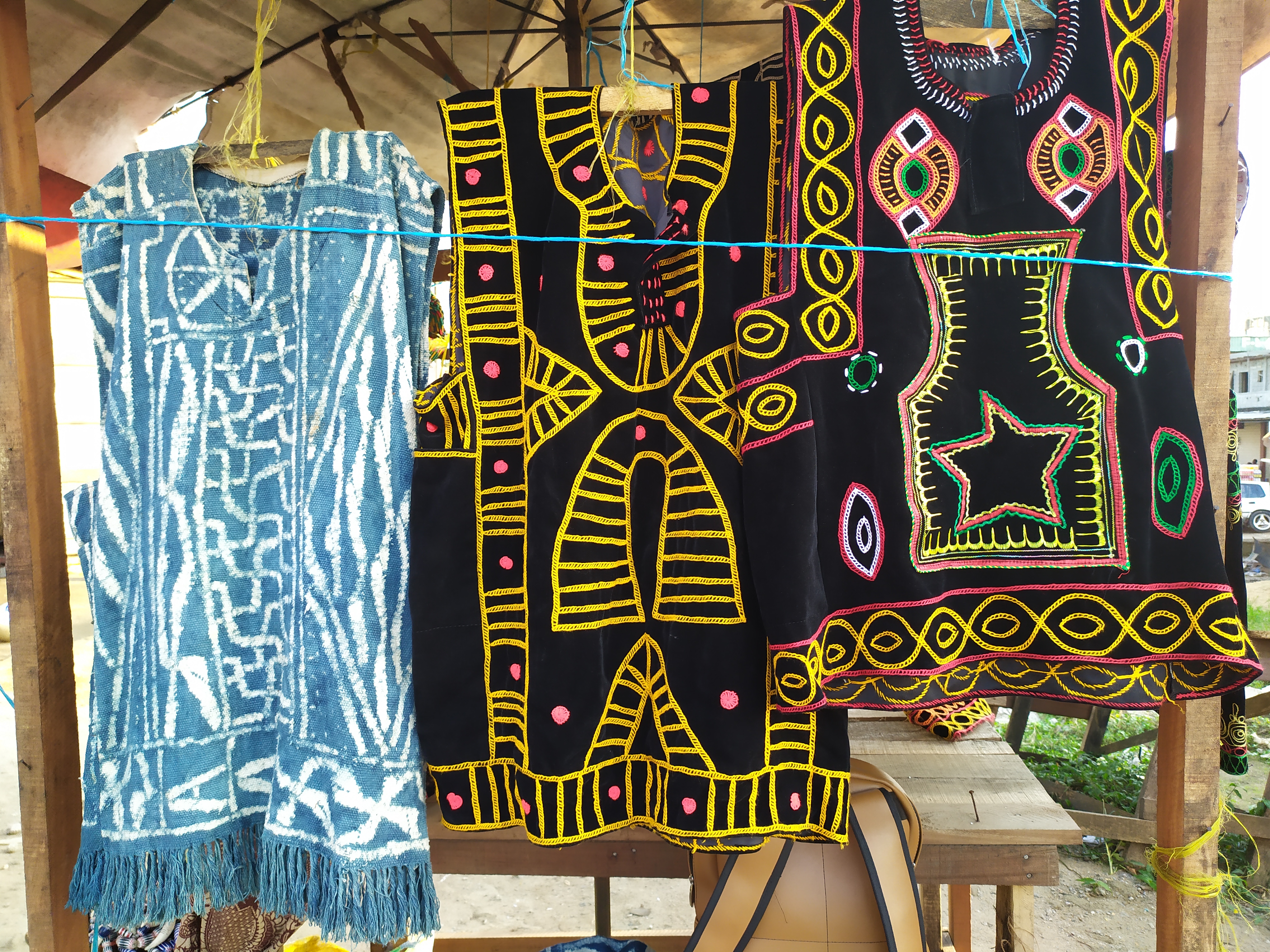 Cameroonian textiles displayed on hangers, with blue-and-white Ndop patterns beside colourful embroidered Toghu garments; archive photograph from 2019